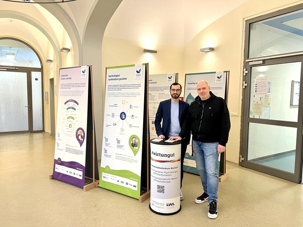 Zwei Männer stehen an einem runden Infostand vor Plakaten und schauen in die Kamera.