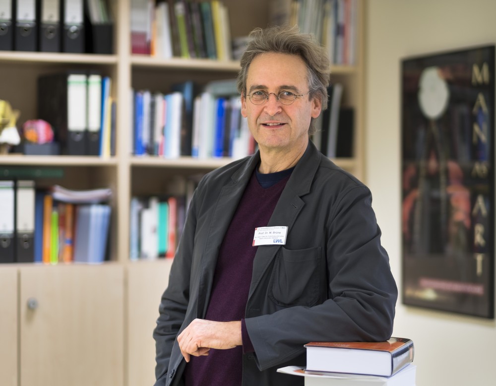Lächelnder Mann mit Brille und grauem Haar, der sich vor Bücherregal auf Büchern abstützt