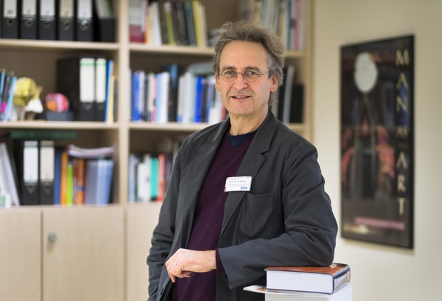 Porträt eines Mannes mit Brille und grauem Haar, der in einem Büro vor einem Bücherregal sitzt. Er trägt ein dunkles Sakko über einem bordeauxroten Pullover und blickt lächelnd in die Kamera.