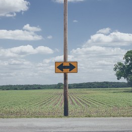 Auf einem breiten Feldweg ist an einem Pfahl ein verwirrendes Schild mit einem Pfeil nach links und einem Pfeil nach rechts