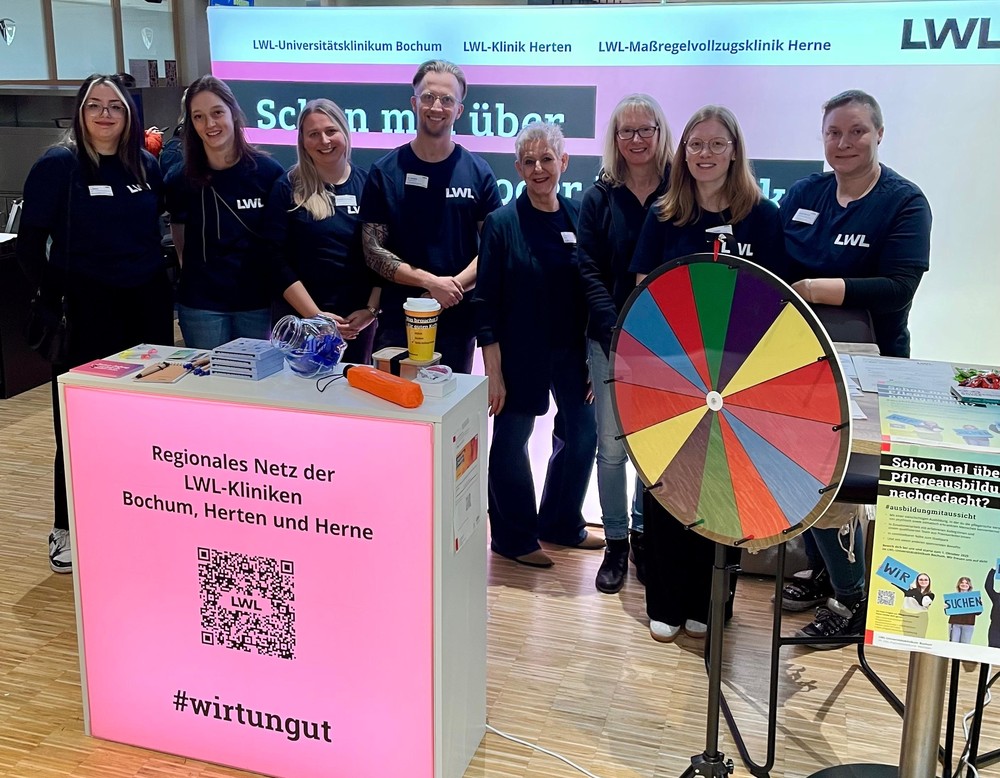 Ein Team der LWL-Kliniken Bochum, Herten und Herne steht gemeinsam an einem Infostand auf der JOBMEDI-Messe. Vor dem Stand steht ein buntes Glücksrad. Alle lächeln in die Kamera.