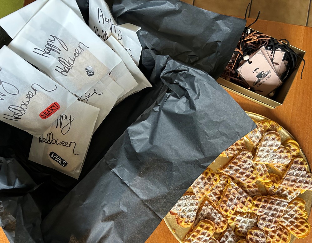 Neben 2 Karton mit Give-Aways ist ein Teller mit Waffelherzen zu sehen, welche mit Puderzucker bestreut sind