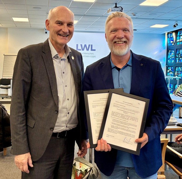 Zwei Männer stehen in einem Konferenzraum vor einem LWL-Logo und halten gerahmte Urkunden in den Händen.