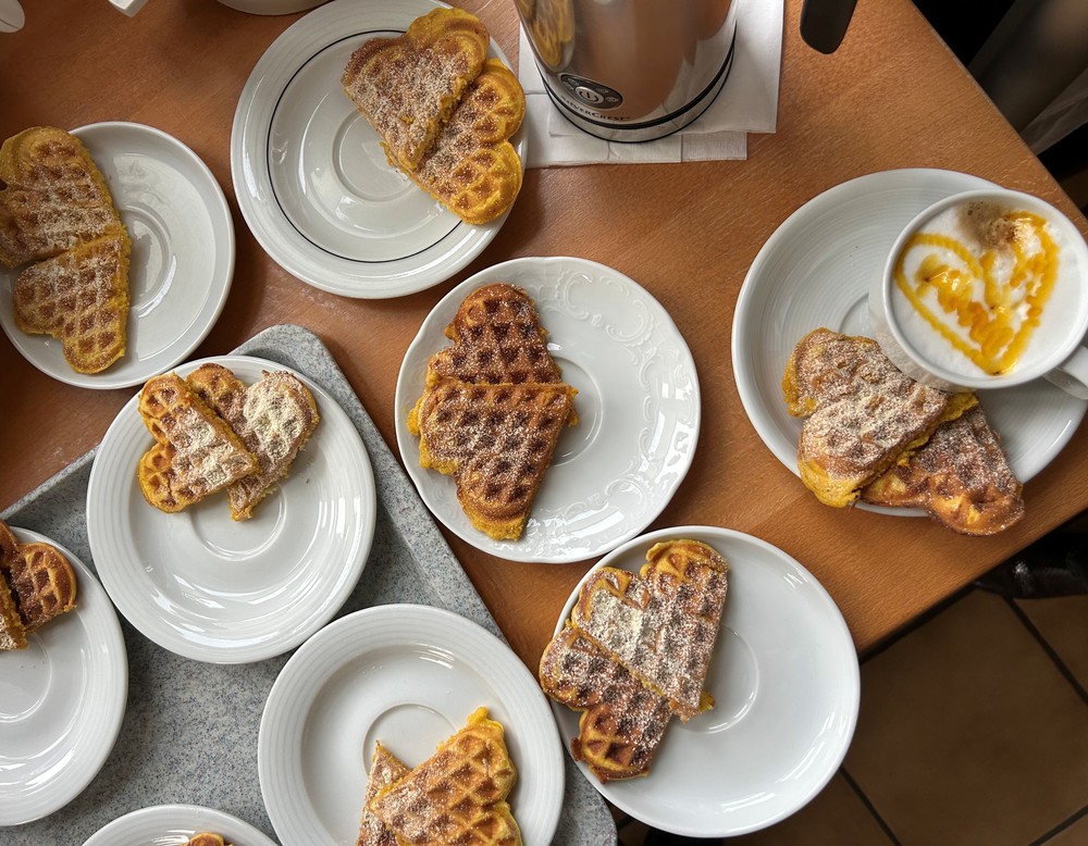Mehrere Unterteller, teils mit Waffelherzen, welche mit Puderzucker bestreut sind, auf einem Tisch von oben fotografiert. Zusätzlich ist eine tasse mit einer Kaffeespezialität mit Milchschaum und einem dekorativen Herz aus Kakaopulver zusehen.