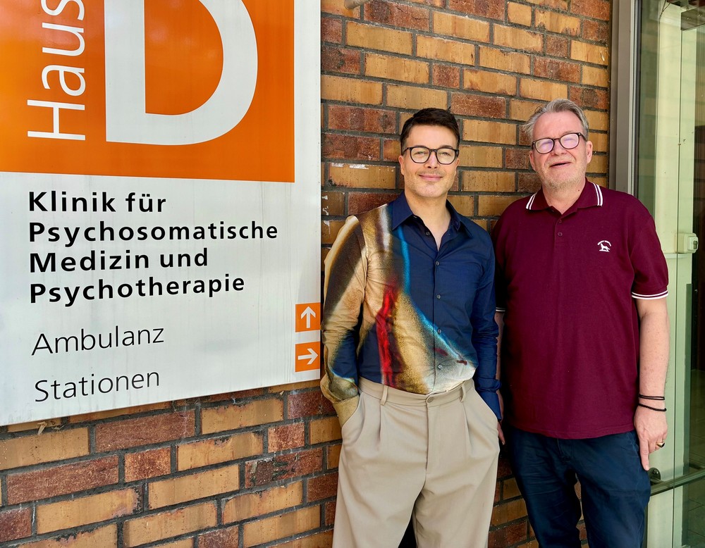 Zwei Personen vor einem Gebäude mit Hinweisschild zur Gebäudebezeichnung
