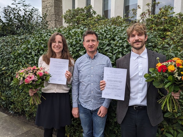 Eine junge Frau und ein junger Mann stehen mit Blumen und Urkunden in der Hand vor einer grünen Hecke und freuen sich. Zwischen ihnen steht ein Mann.