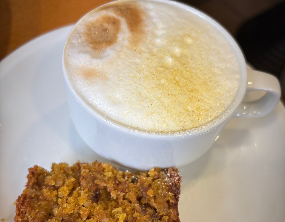 Kaffeespezialität mit Milchschauf in einer weißen Tasse auf einem Unterteller. Zusätzlich ist ein kleines Stück Gebäck auf dem Unterteller platziert